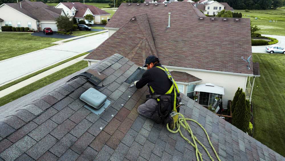 A man is repairing the roof of a house under a clear blue sky.